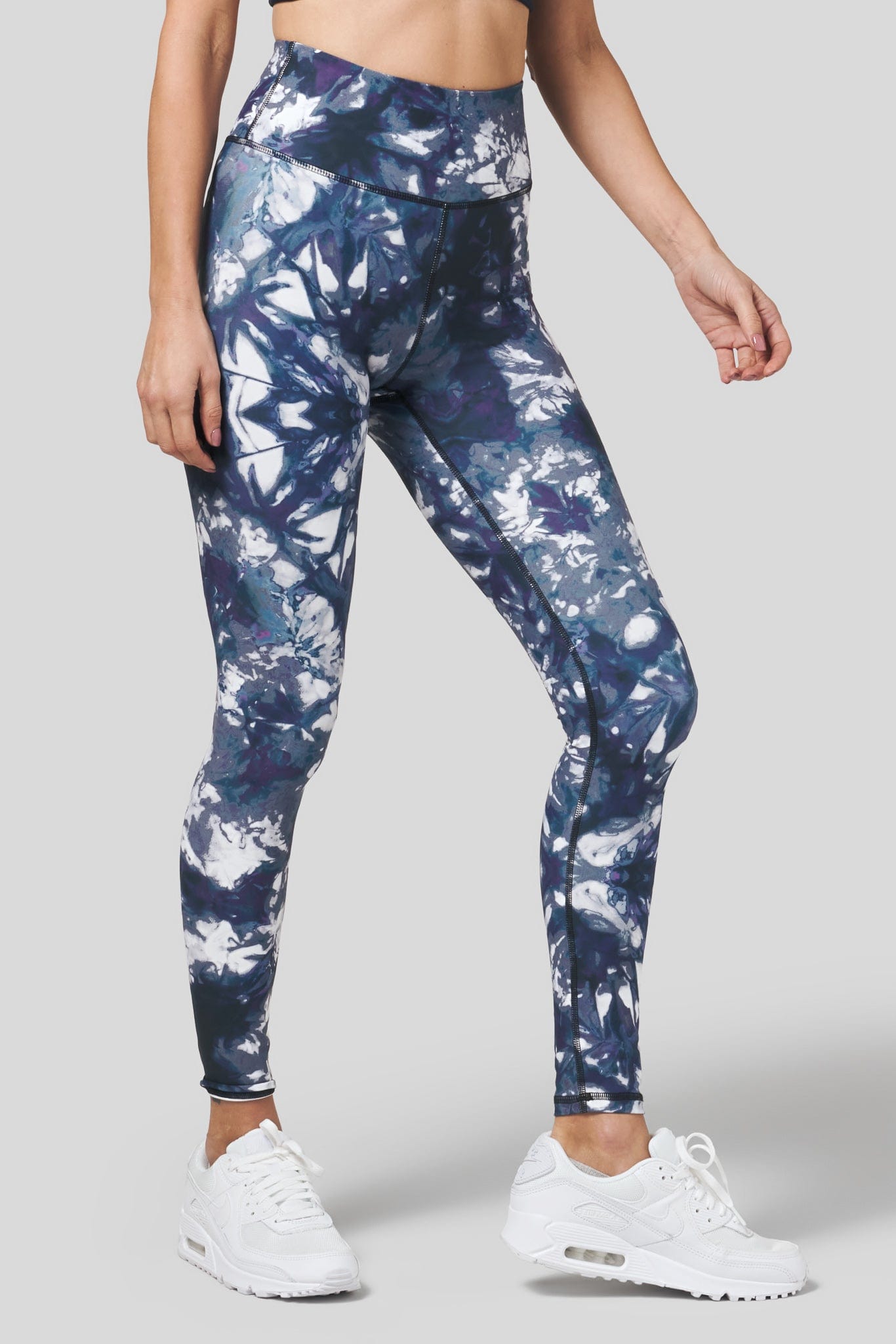 A woman wears a pair of non-compressive leggings in a tie dye print with white Nike Air Max 90s.