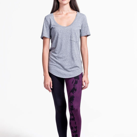 A woman with brown hair models a black sports bra and leggings. The right pant leg of the leggings is black, while the other is tie-dyed in plum and black.