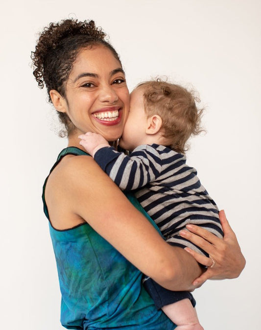 Marimba holds her baby boy and smiles at the camera. She wears a tie-dye tank top and he wears a striped onesie.
