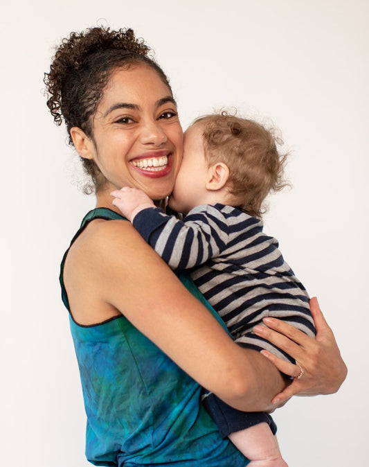 Marimba holds her baby boy and smiles at the camera. She wears a tie-dye tank top and he wears a striped onesie.