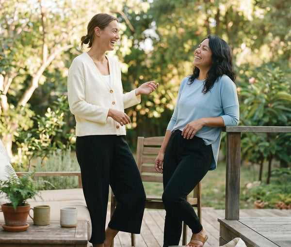 Two women chatting and laughing on a patio, styled in comfortable DAUB shirt, cardigan and casual pants.