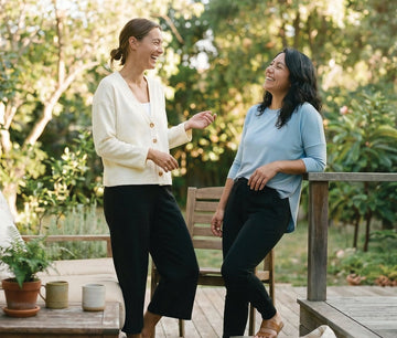 Two women chatting and laughing on a patio, styled in comfortable DAUB shirt, cardigan and casual pants.