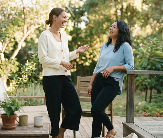 Two women chatting and laughing on a patio, styled in comfortable DAUB shirt, cardigan and casual pants.