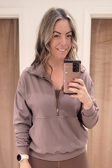 A blonde woman wears a light brown half zipped sweatshirt with ribbed leggings and a sports bra. Each piece is made in Canada.