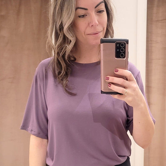A woman wears a purple tee shirt with a loose sleeve and loose body. It's tucked into a pair of black sweatpants.