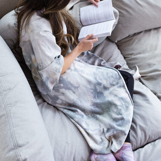 A woman sits on a grey couch and reads a book while curled up in a tie-dyed robe.