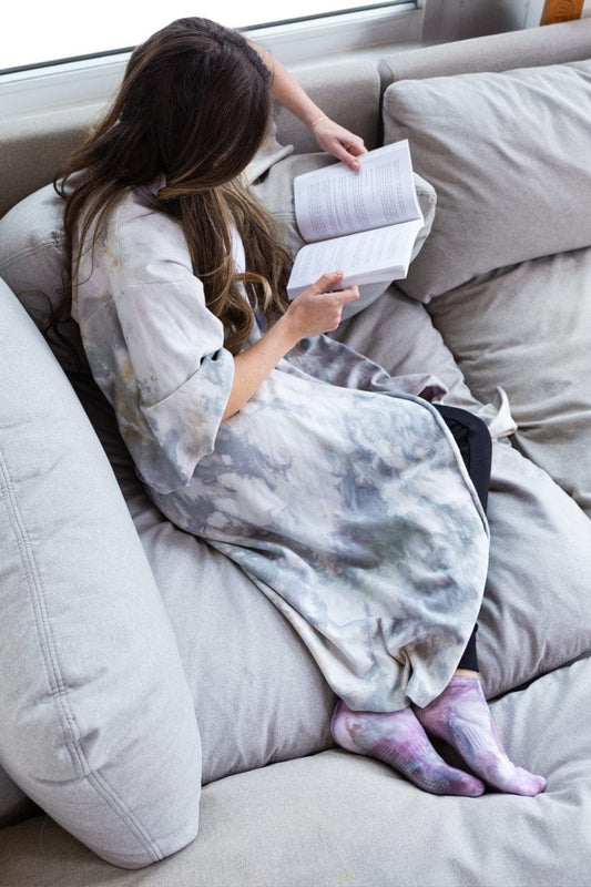 A woman sits on a grey couch and reads a book while curled up in a tie-dyed robe.