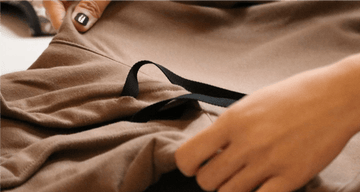 A person with painted nails adjusts a brown fabric, pulling a black ribbon through a seam. The focus is on the fabric and hands. By Daub & Design.