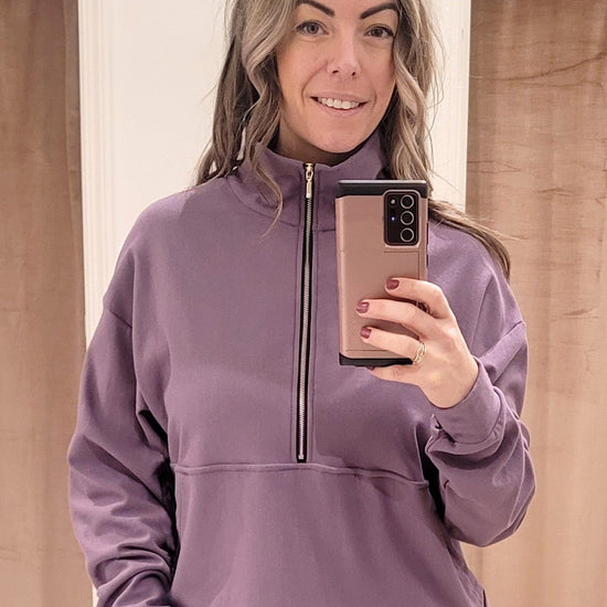 A blonde woman wears a light purple crewneck sweatshirt made from bamboo cotton fleece. The sweatshirt has a gold zipper that opens halfway down the front with a kangaroo pouch.