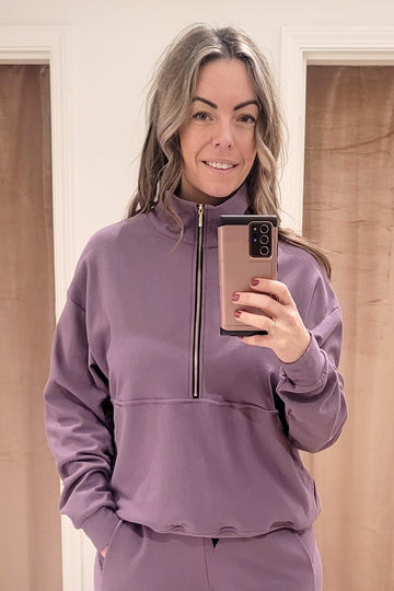 A blonde woman wears a light purple crewneck sweatshirt made from bamboo cotton fleece. The sweatshirt has a gold zipper that opens halfway down the front with a kangaroo pouch.