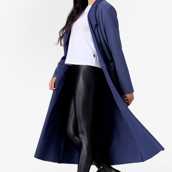 A woman walks with an open front long jacket that reaches her mid calf. The jacket is blue and she has a white tee shirt and liquid black leggings