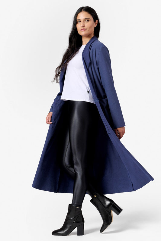 A woman walks with an open front long jacket that reaches her mid calf. The jacket is blue and she has a white tee shirt and liquid black leggings