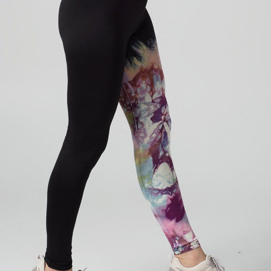 A woman with red hair models a black sports bra and leggings. The right pant leg of the leggings is black, while the other is tie-dyed bright and purple colours.