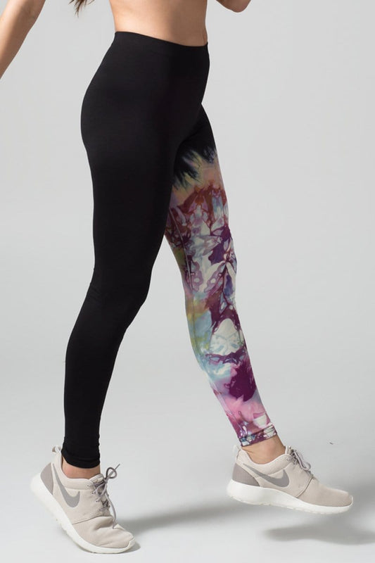 A woman with red hair models a black sports bra and leggings. The right pant leg of the leggings is black, while the other is tie-dyed bright and purple colours.