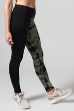 A woman with brown hair models a black sports bra, leggings, and grey sneakers. The right pant leg of the leggings is black, while the other is tie-dyed in gold and black.