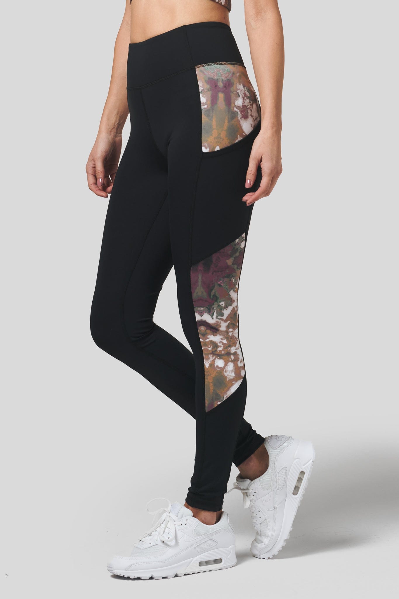 A woman with white Nike Air Max sneakers wears a pair of DAUB pocket leggings in black with a side panel in earthy tie-dye.