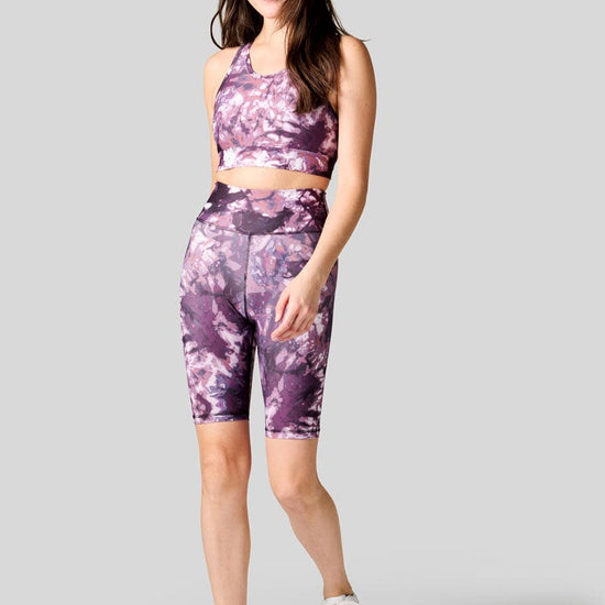 A dark haired woman wears a high neck sports bra in tie dye with a pair of high waisted Bike Shorts