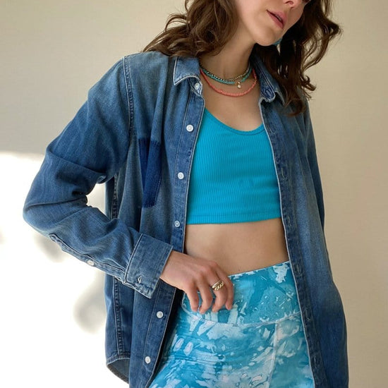 A woman wears a blue ribbed reversible top with blue tie-dye bike shorts and a denim button down shirt.