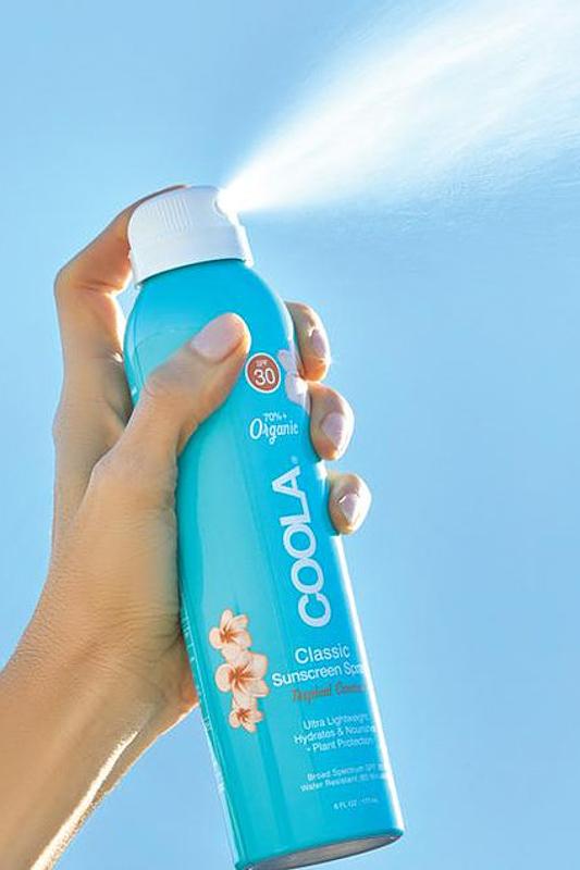 Hand spraying suncreen from blue spray bottle with "Coola" written across the front