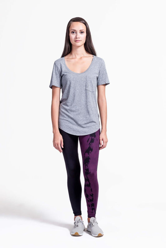 A woman with brown hair models a black sports bra and leggings. The right pant leg of the leggings is black, while the other is tie-dyed in plum and black.